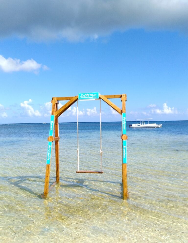 A wooden swing set with turquoise hashtags and text stands in shallow, clear waters on Roatan, Honduras, with a boat and the ocean visible in the background under a partly cloudy sky.