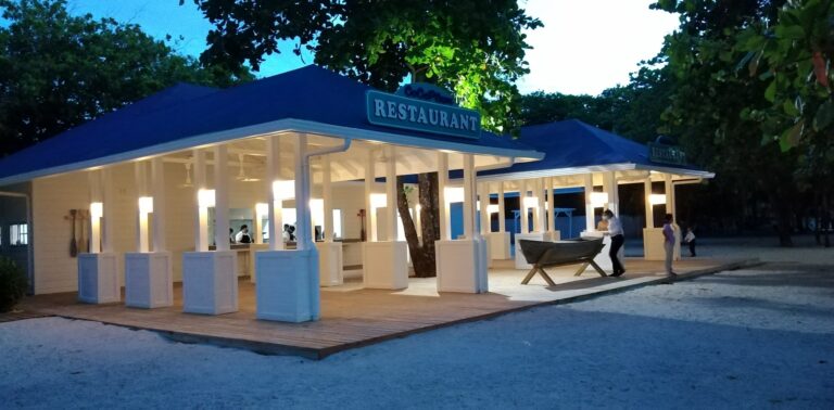 A brightly lit white restaurant with an open-air design is set among trees in Roatan, Honduras, during dusk, with staff and guests visible near the entrance.