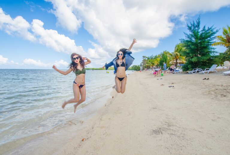 Two women in bikinis and sunglasses jump and pose on a sandy beach with clear water, palm trees, and lounge chairs along the shore, with a blue sky and white clouds over Roatan in Honduras.