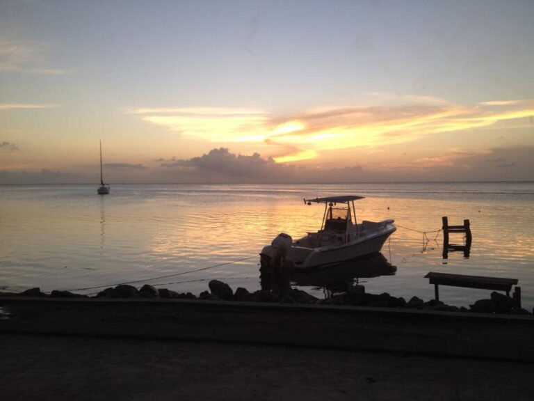 A small boat is anchored near the rocky shoreline at sunset in Roatan, Honduras, with calm waters, a distant sailboat, and a partially submerged wooden dock visible in the background.