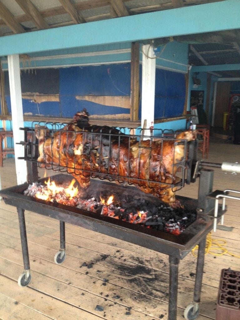 A roasted pig is cooking on a spit over an open flame inside a rustic, open-air structure in Roatan, Honduras, with wooden flooring and blue-painted walls.