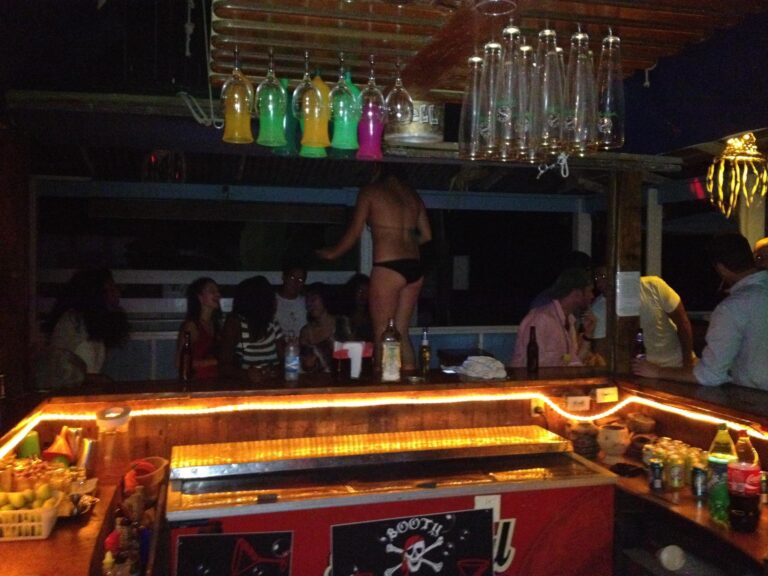A bar area in Roatan, Honduras, features hanging colorful shot glasses and tall glasses, with a group of people enjoying drinks beneath a wooden ceiling and warm orange lighting.