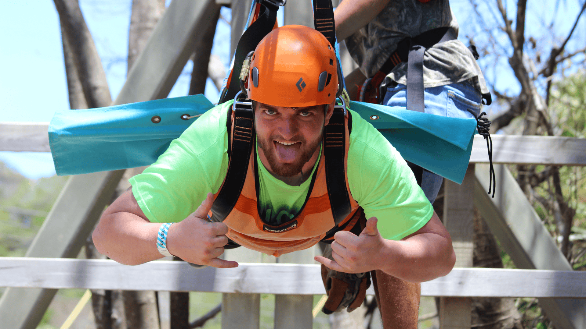 A man wearing an orange helmet and bright green shirt gives a thumbs-up while participating in an outdoor climbing or adventure activity in Roatan, Honduras.