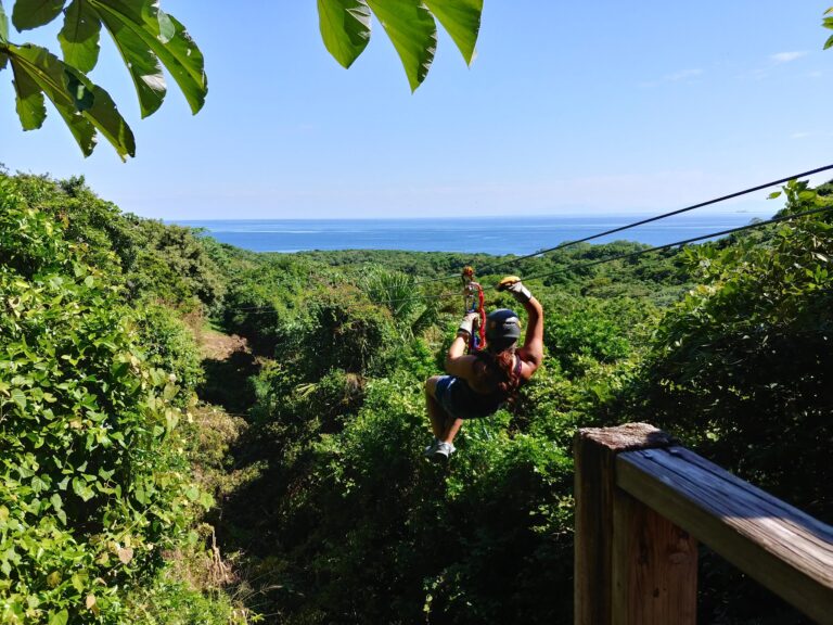 A person wearing a helmet and gloves is ziplining over lush green foliage with a view of the ocean and distant ships in Roatan, Honduras.