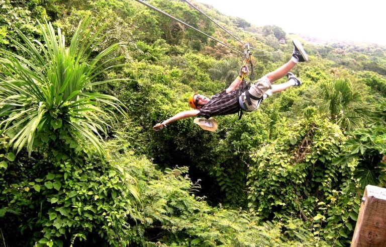 A person zip-lining through lush green vegetation on Roatan in Honduras, wearing a helmet, harness, and sunglasses, with a tropical forest and hillside in the background.