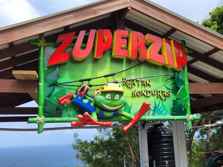 A colorful wooden sign reading "Zuper Kido" with a cartoon frog, red chili peppers, and other playful decorations, mounted on a wooden structure overlooking the ocean in Roatan, Honduras.