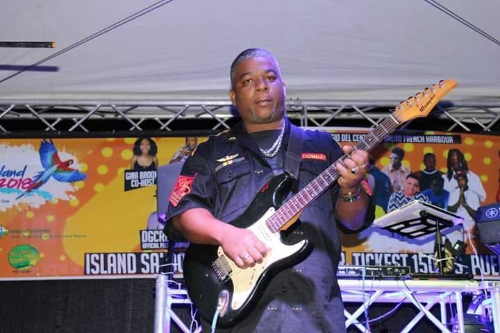 A man wearing a black uniform and silver chain plays an electric guitar on stage with a colorful island-themed banner in the background, promoting events in Roatan, Honduras.