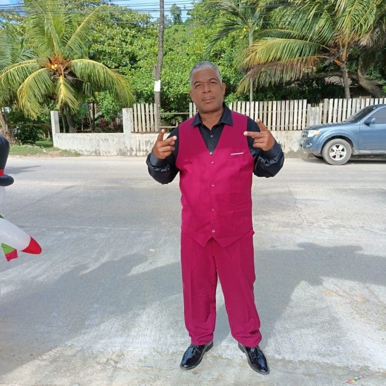 A man in a black shirt and bright pink vest and pants poses on a street in Roatan, Honduras, pointing with both hands, with tropical palm trees, a wooden fence, and parked cars visible in the background.