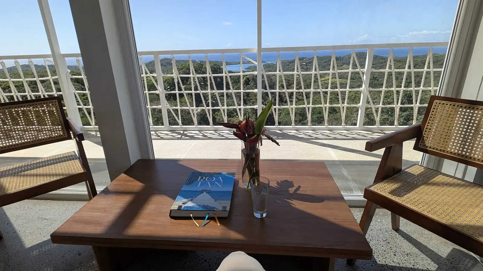 Interior of a modern balcony in Roatan, Honduras, with a wooden table, two chairs, and a glass door opening to a scenic view of lush green hills and the Caribbean Sea.