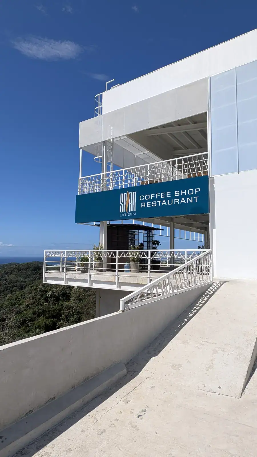 Modern white building with a blue sign reading 'Spirit Coffee Shop Restaurant' on a balcony overlooking lush green islands in Roatan, Honduras under a clear blue sky.