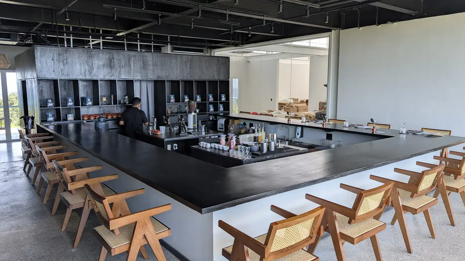 Modern upscale bar with a black marble countertop, surrounded by wooden chairs with woven seats, located in a bright, open space on Roatan in Honduras, featuring a partially visible kitchen and a bartender preparing drinks.