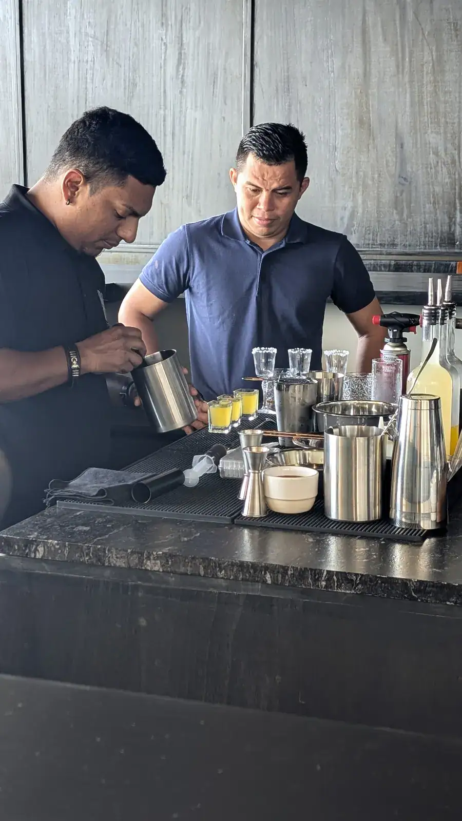 Two bartenders preparing cocktails behind a black granite bar counter in Roatan, Bay Islands, Honduras, with various bar tools and glasses visible.
