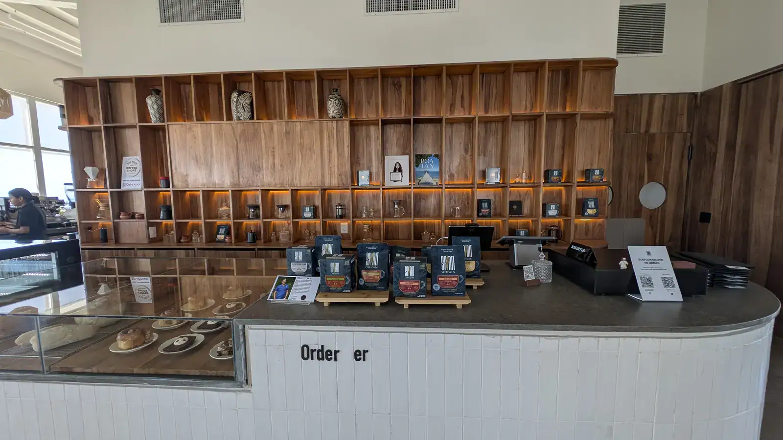 Interior view of a modern coffee shop in Roatan, Bay Islands, Honduras, featuring a wooden shelving unit with decorative vases and coffee-related items behind the counter, and various packaged coffee products displayed on the countertop.