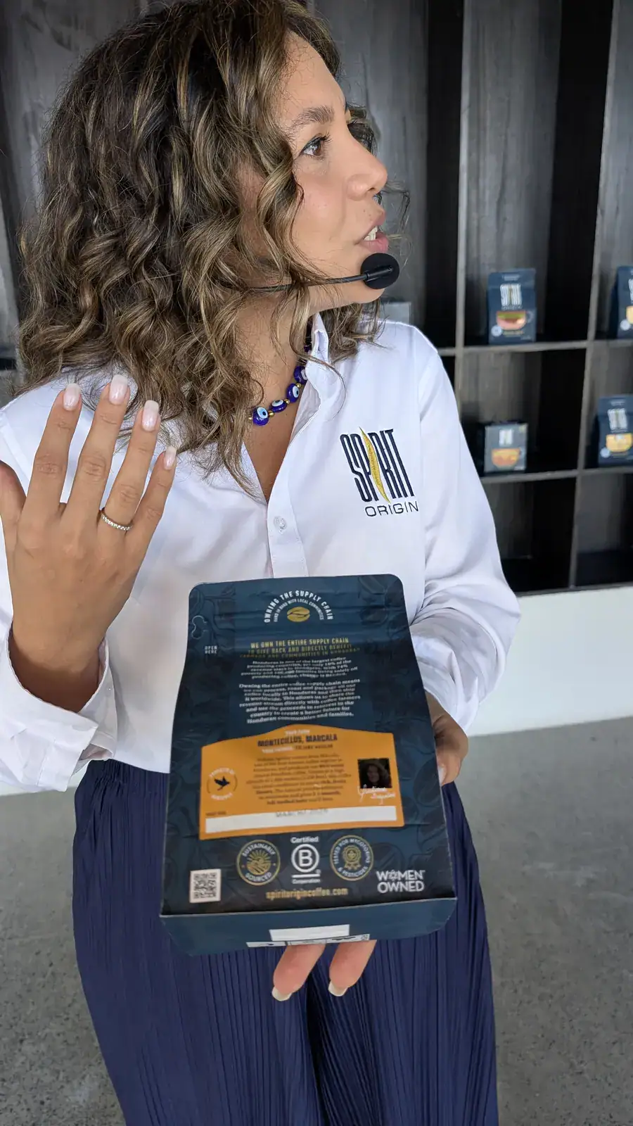 A woman in a white shirt with Roatan Origin logo explaining a product at a business event in Roatan, Honduras, with a display of packages behind her.