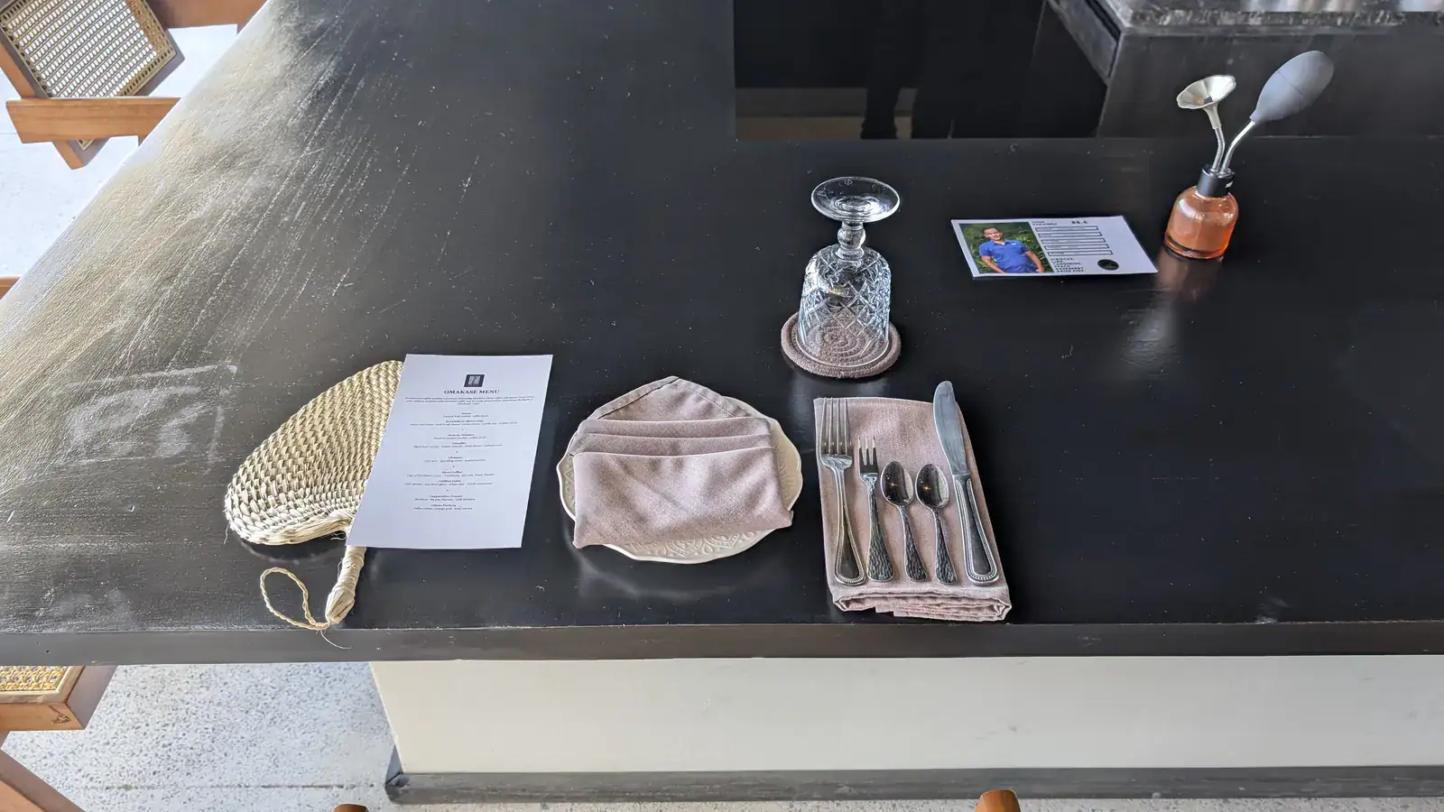 A neatly set dining table at a restaurant in Roatan, Honduras, featuring utensils, a wine glass, a folded napkin on a ceramic plate, a menu, and a small decorative bottle with utensils inside.