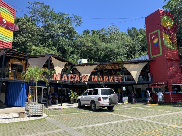 Macao Market, a vibrant shopping and dining area in Roatan, Honduras, features colorful shipping container structures and lively outdoor seating with people shopping and dining under a clear blue sky.