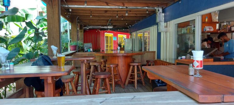 The image shows an open-air restaurant or bar with wooden tables and stools, colorful walls including red, yellow, and blue, and lush green tropical plants in the background, located in Roatan, Honduras.