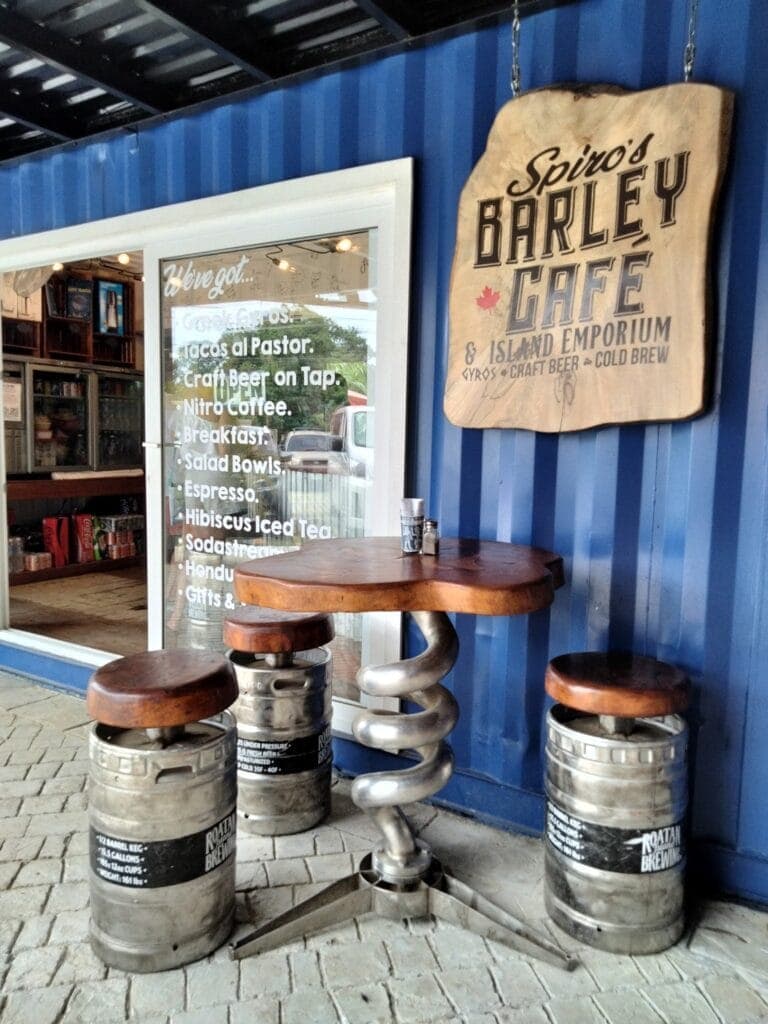 A seating area features three barstools made from repurposed beer kegs with wooden seats and metal spring bases outside a blue-walled establishment in Roatan, Honduras.