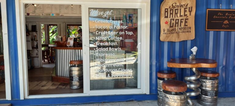 A blue wooden storefront on Roatan, Honduras, features a glass door with a menu listing Greek gyros, tacos al pastor, craft beer on tap, and other items, alongside a seating area with stools made from kegs and a large wooden sign for Spino's Barley Cafe.