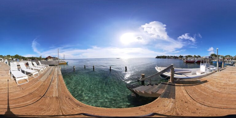 A wooden dock along the shoreline in Roatan, Honduras, features white lounge chairs, several boats docked nearby, and a clear blue sky with the sun reflecting on the calm water.