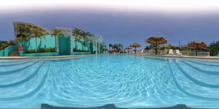 An outdoor swimming pool at a resort in Roatan, Honduras, features clear blue water, surrounded by white lounge chairs and thatched umbrellas, with colorful tropical landscaping and buildings in the background.