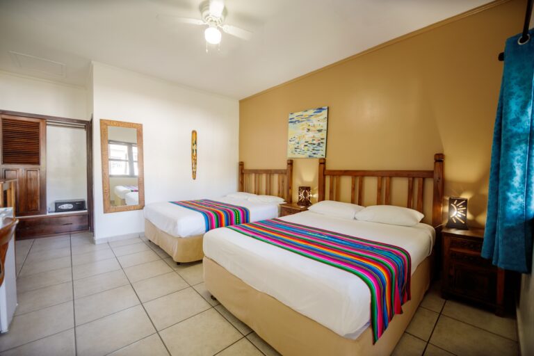 Two neatly made beds with colorful striped blankets are shown in a bright, tropical-style hotel room on Roatan, Honduras, featuring wooden furniture, tropical decor, and a window with blue patterned curtains.