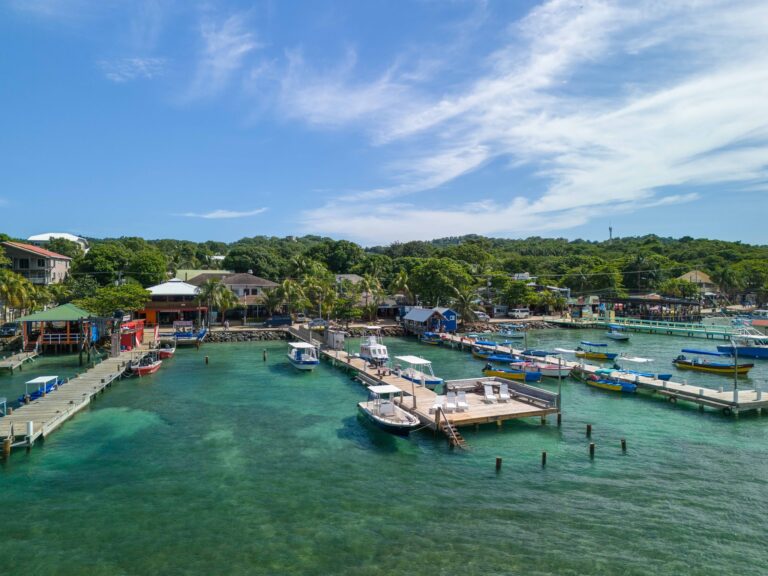 A picturesque harbor in Roatan, Honduras, features turquoise waters, numerous small boats docked at piers, and lush green hills dotted with residential buildings and tropical trees under a bright blue sky.
