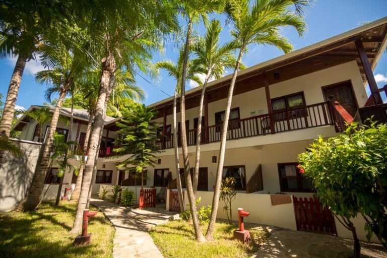 A multi-story building with wooden balconies surrounded by tall palm trees and lush tropical vegetation under a bright blue sky in Roatan, Honduras.