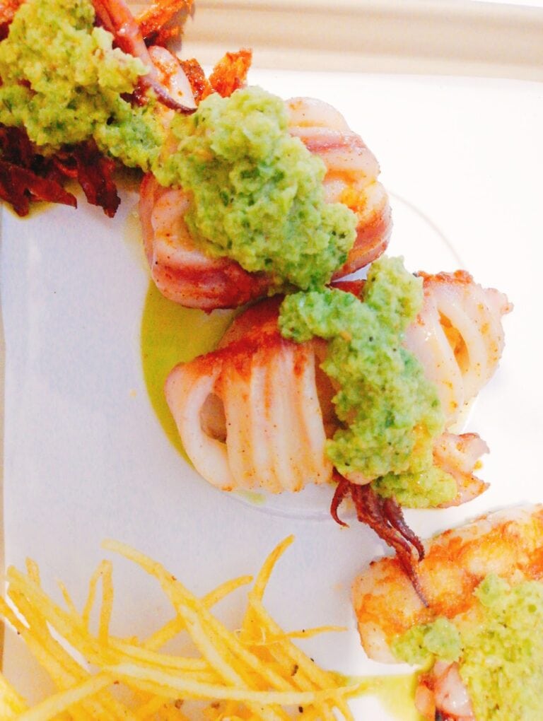 Fresh grilled shrimp topped with guacamole and served with crispy fried potatoes on a white plate, with a colorful background indicating a meal setting in Roatan, Honduras.