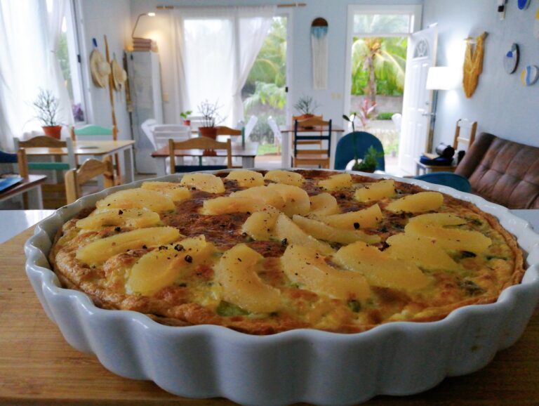 A pineapple-topped pizza in a white ceramic dish is set on a wooden table inside a bright, airy home in Roatan, Honduras, with windows revealing lush green tropical foliage outside.
