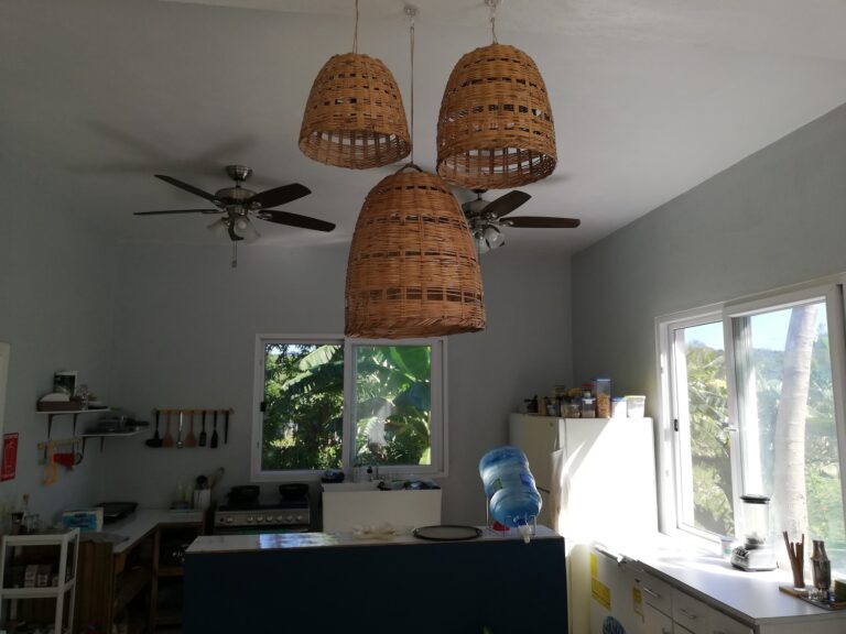 A bright, airy kitchen in Roatan, Honduras, features white walls, large windows with views of lush greenery, woven wicker pendant lights hanging from the ceiling, and various kitchen appliances and utensils organized on shelves and counters.