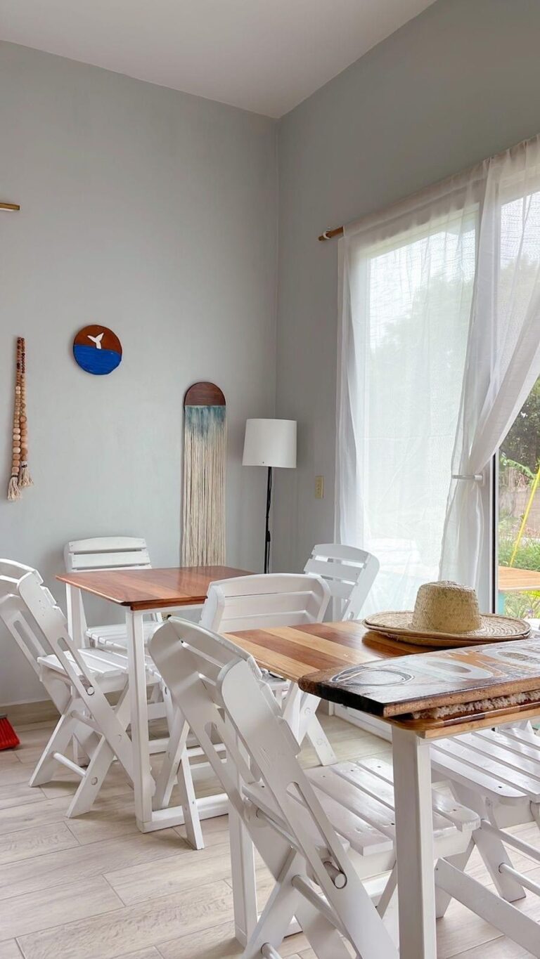 A bright dining area in Roatan, Honduras, features white wooden chairs around tables with natural wood tops, a large window with sheer curtains allowing natural light, and seaside-themed wall decorations including a whale tail and ocean artwork.