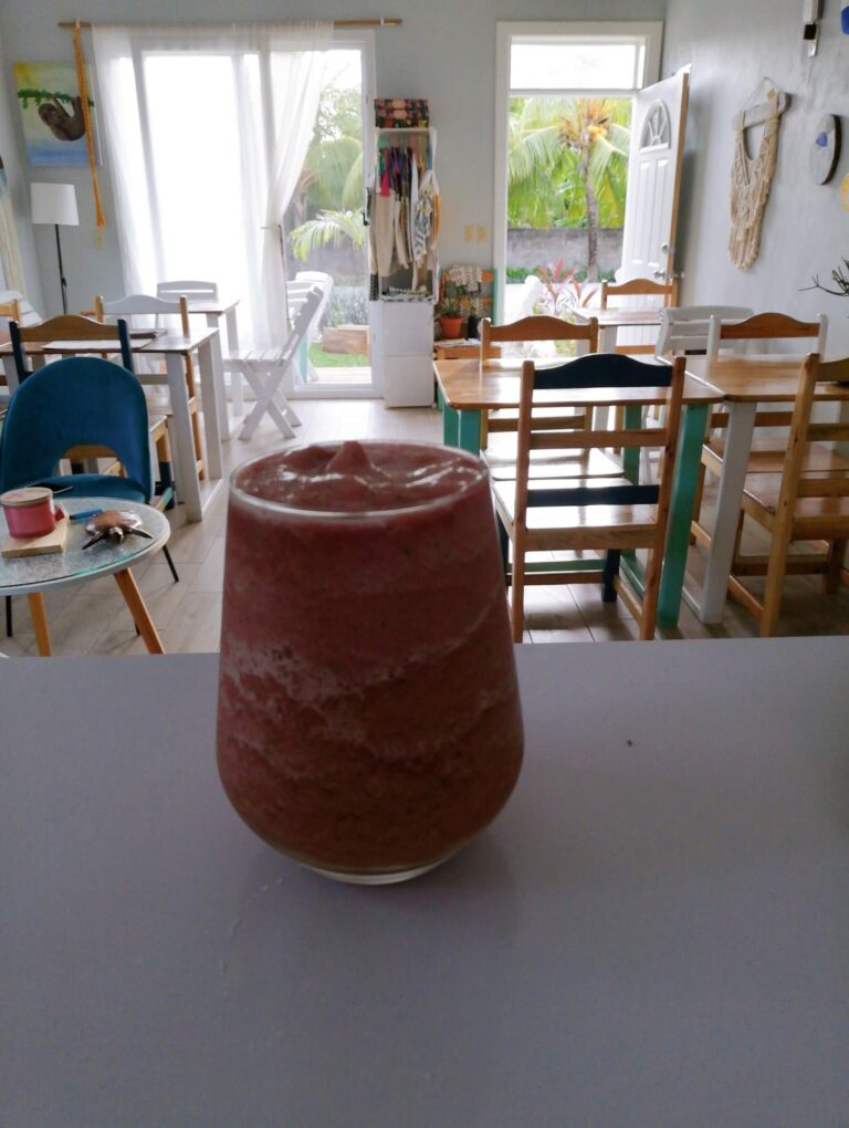 A ceramic vase is placed on a white surface inside a room with wooden tables and chairs, large windows with white curtains, and tropical plants visible outside in Roatan, Honduras.