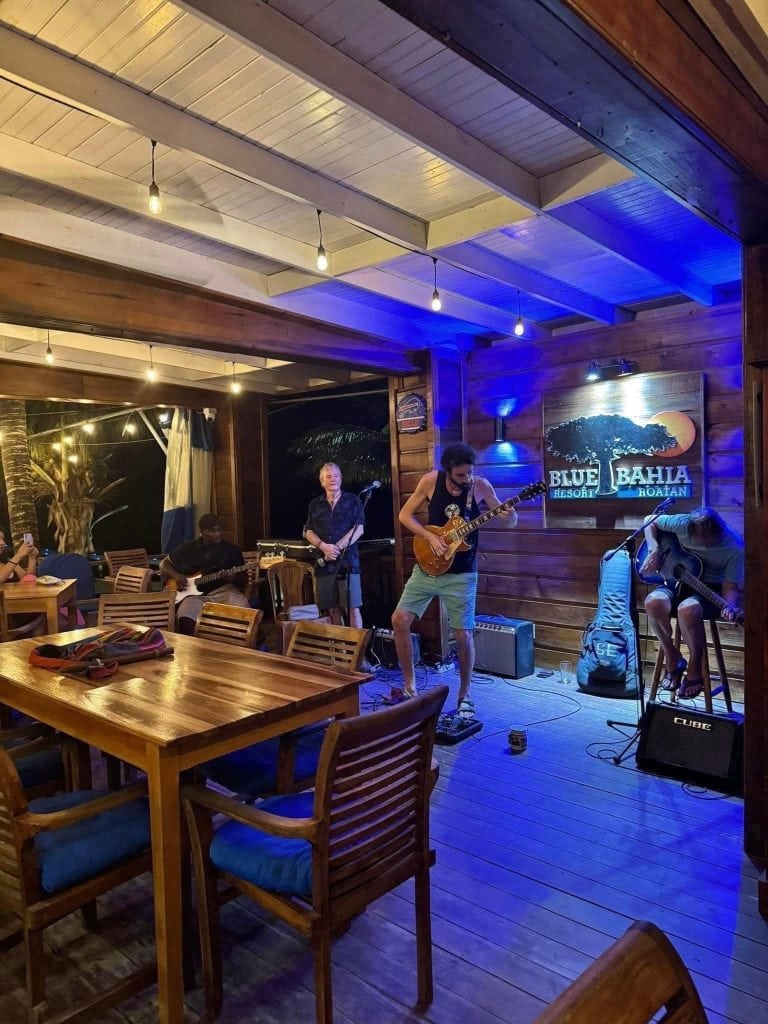 A live music performance takes place inside a wooden restaurant or bar in Roatan, Honduras, featuring musicians playing guitars with an audience seated at tables and a large illuminated sign reading "Blue Bahia Resort Roatan.