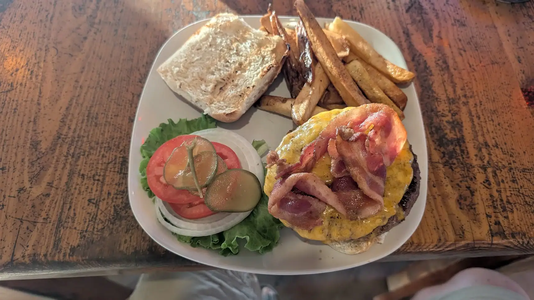 A plate of Honduran and Roatan-style food including a cheeseburger with bacon, French fries, sliced tomatoes, cucumbers, onions, and a slice of bread on a wooden table.