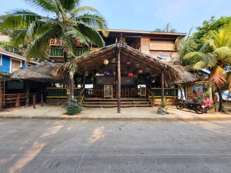 A two-story wooden bar or restaurant with thatched roofing and tropical decor, situated among palm trees on Roatan, Honduras, with colorful signage and a mural featuring a marlin fish.