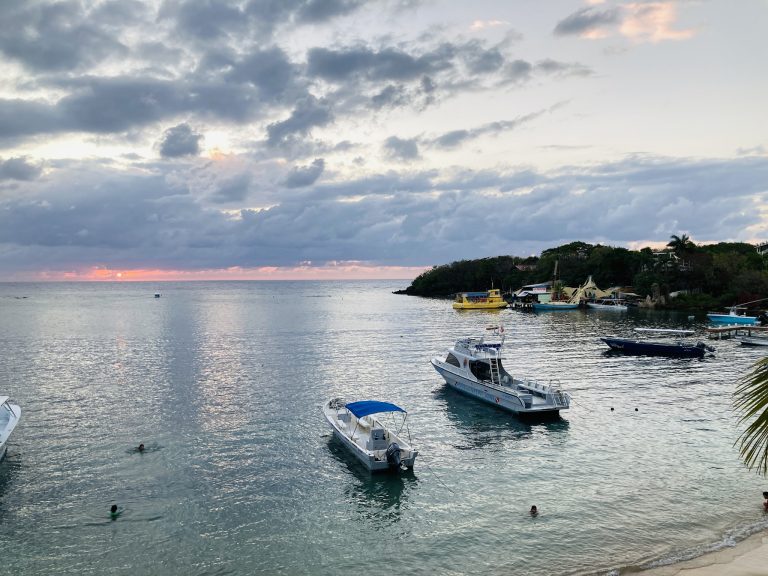 A view of Roatan's coastline in Honduras at sunset shows boats docked near the shore, people swimming in the water, and lush green hills with buildings and palm trees under a partially cloudy sky.