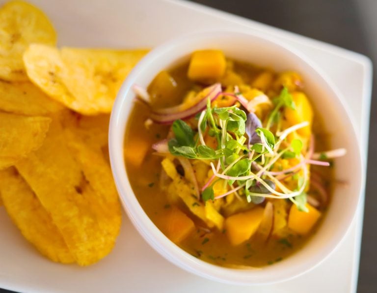 A bowl of Honduran chicken or fish soup garnished with microgreens, accompanied by tortilla chips, set on a white surface in Roatan, Bay Islands.