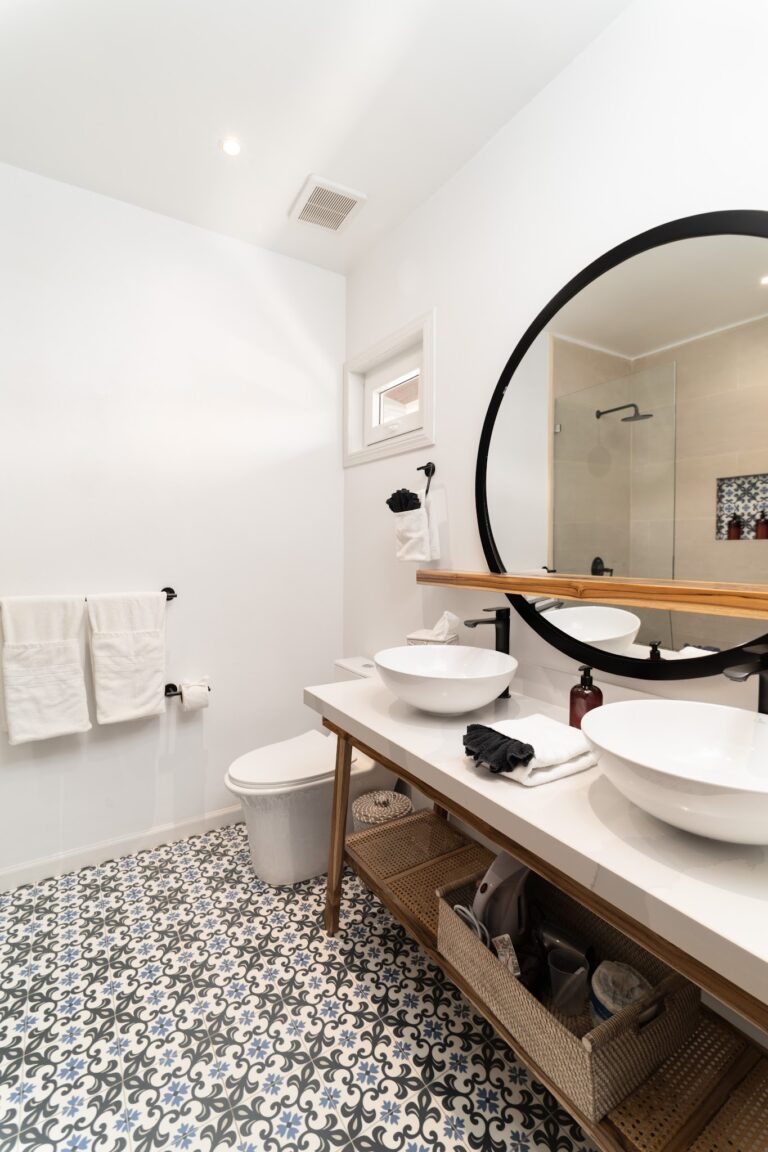 A modern bathroom in Roatan, Honduras, features a white double sink vanity with vessel sinks, a large circular mirror, patterned black and white floor tiles, and a wicker storage shelf beneath the countertop.