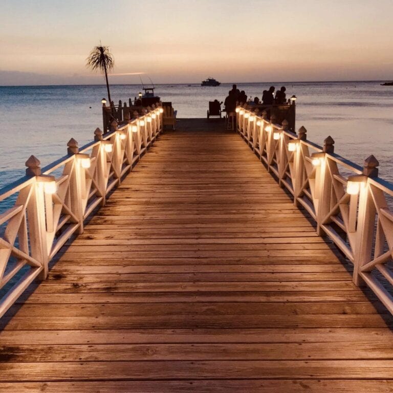 A wooden pier with illuminated lamps leading to a dining area on the water at sunset in Roatan, Honduras, with silhouettes of people and a boat in the distance.