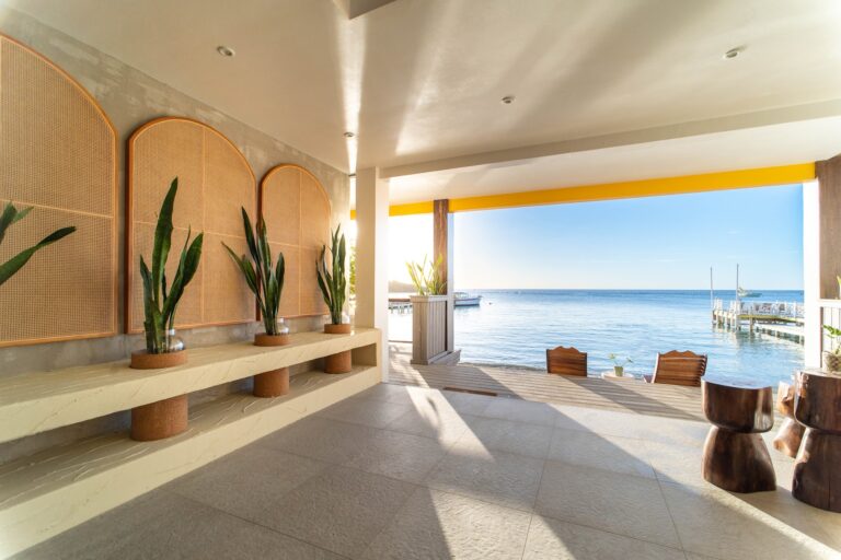 A modern indoor seating area with a view of the ocean, featuring wooden chairs, potted plants, and a partially open wall revealing boats and a pier in Roatan, Honduras.