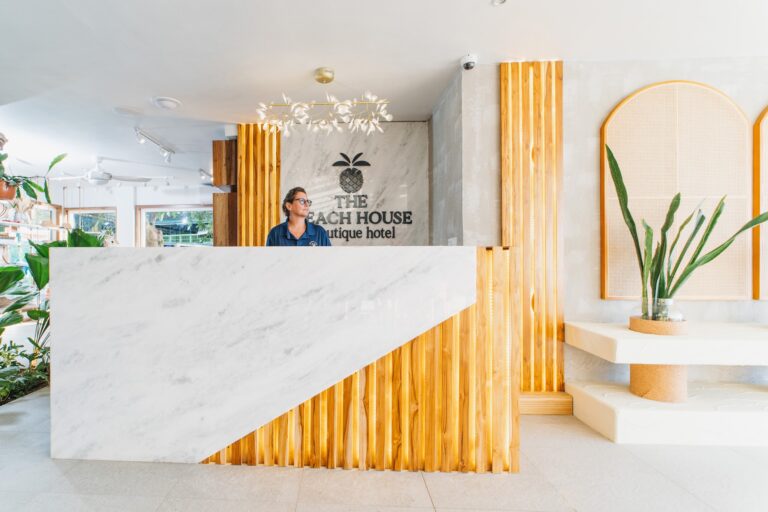 The reception area of The Beach House Boutique Hotel in Roatan, Honduras features a white marble front desk with wooden paneling, a decorative pineapple sign on the wall, and a modern chandelier with white floral accents, accompanied by lush green plants.