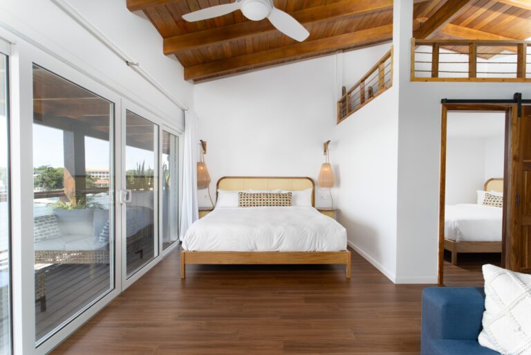 A modern bedroom with a wooden ceiling and floor, featuring a bed with a woven headboard and decorative pillows, large sliding glass doors leading to a balcony with outdoor seating, and partial views of other beds with a wooden railing, located on Roatan, Honduras.