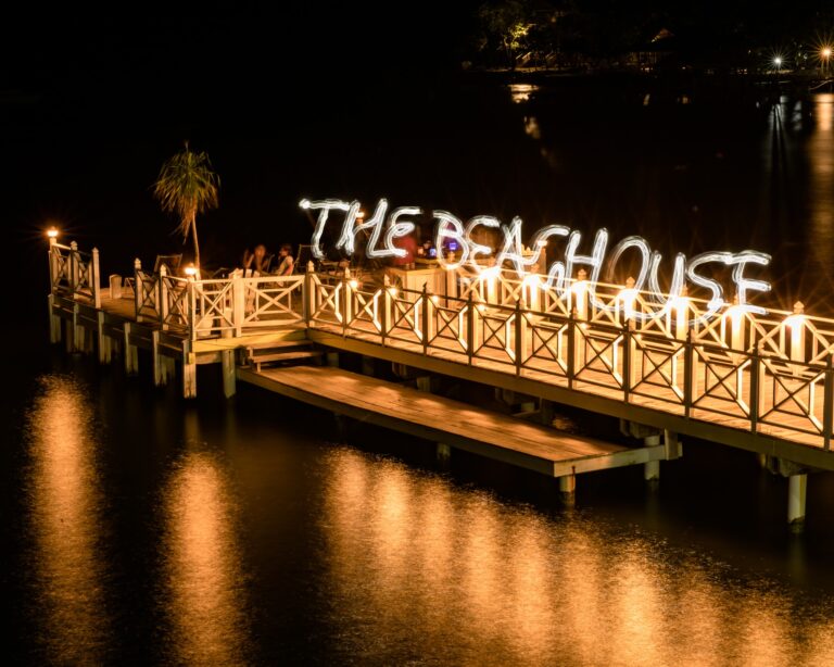 People sitting on a wooden pier illuminated by string lights with a large neon sign saying "The Beach House" over Roatan, Honduras, at night, reflecting on calm water.