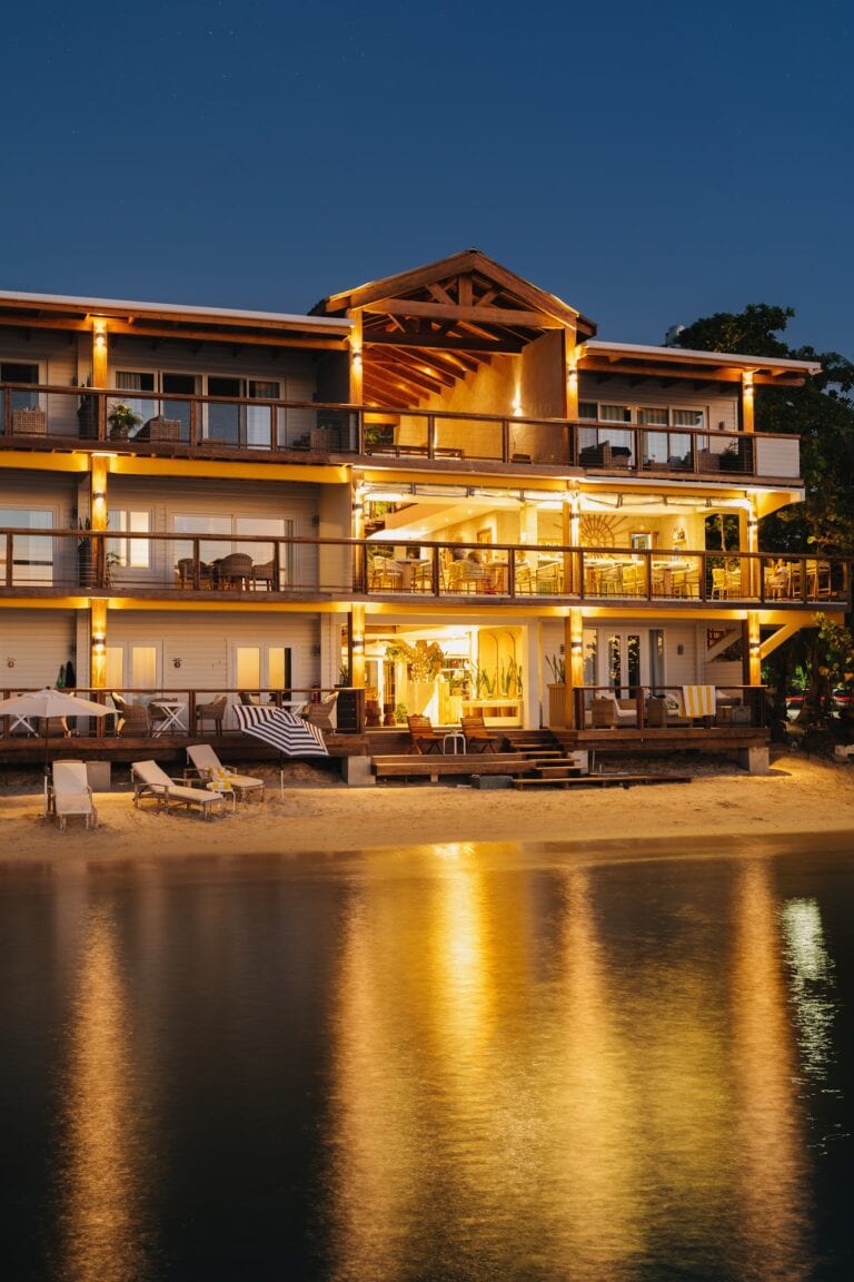 A three-story beachfront hotel in Roatan, Honduras, with outdoor seating on all levels, illuminated by warm lights at night, reflecting on the calm water in front.
