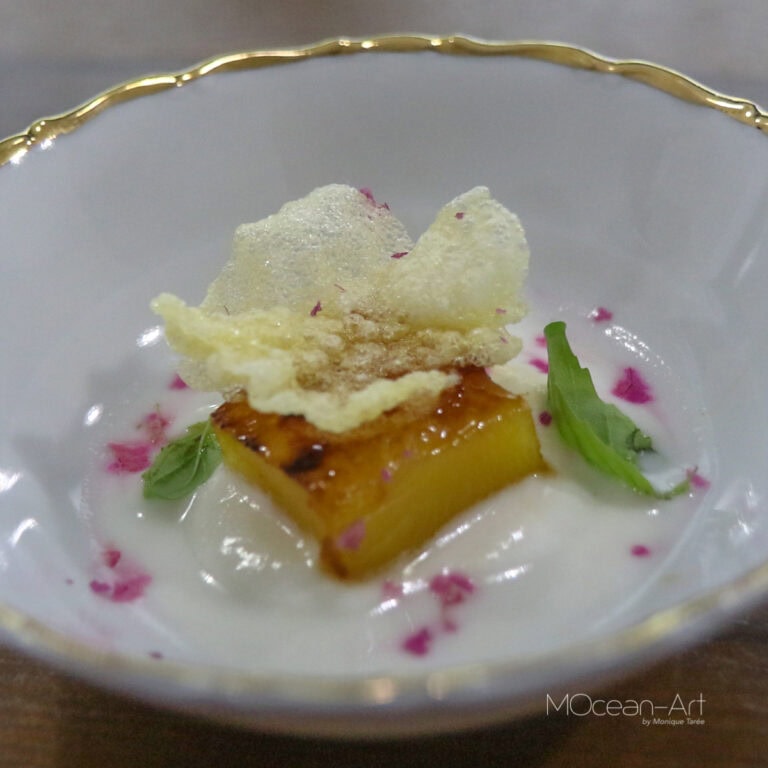 A plated dessert featuring a caramelized pineapple chunk topped with a crispy tuile, garnished with small green leaves and pink edible flower petals, served in an elegant glass bowl on Roatan, Honduras.