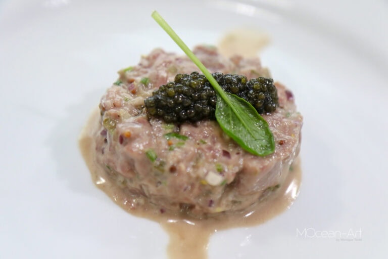 A serving of tuna tartare topped with black caviar and a green leaf garnish is presented on a white plate, with a blurred background suggesting a casual dining setting in Roatan, Honduras.