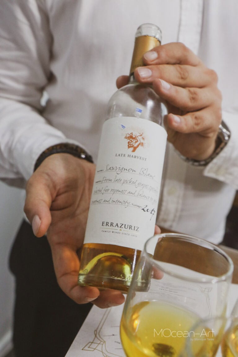 A person holds a bottle of wine labeled "Late Harvest" and "Errazuriz" in front of a glass filled with a golden-colored beverage, with a setting that suggests a wine tasting event on Roatan, Honduras.