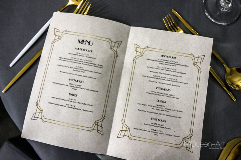 A folded printed menu with gold decorative borders is displayed on a dark table setting next to gold and white cutlery and a wine glass, situated in a restaurant in Roatan, Honduras.