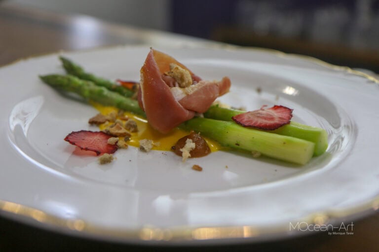 A plate of gourmet food with asparagus, prosciutto, strawberry slices, crumbled cheese, and a drizzle of sauce is presented on an elegant white dish with a gold-rimmed edge, set against a blurred background, in Roatan, Honduras.
